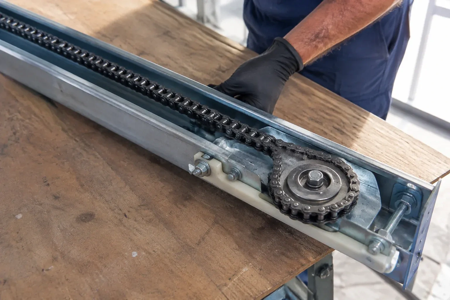 Gloved hand resting on a steel opener rail laid on a wooden bench, showing a drive chain looped around a sprocket wheel. — Chain-Drive Opener Repair, Peterborough
